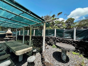 A covered outdoor seating area with stone benches and a table, surrounded by a stone wall and greenery.