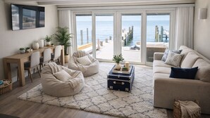 En moderne stue med sofa, to sækkestole, et sofabord med et skrin, et spiseområde med bord og stole, og en stor skydedør, der fører ud til en balkon med havudsigt.
