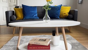 Un salon moderne avec une table ronde blanche, un vase rempli de fleurs jaunes, un livre intitulé « Vivre simplement à l’ère moderne » et un canapé agrémenté de coussins bleus et jaunes.