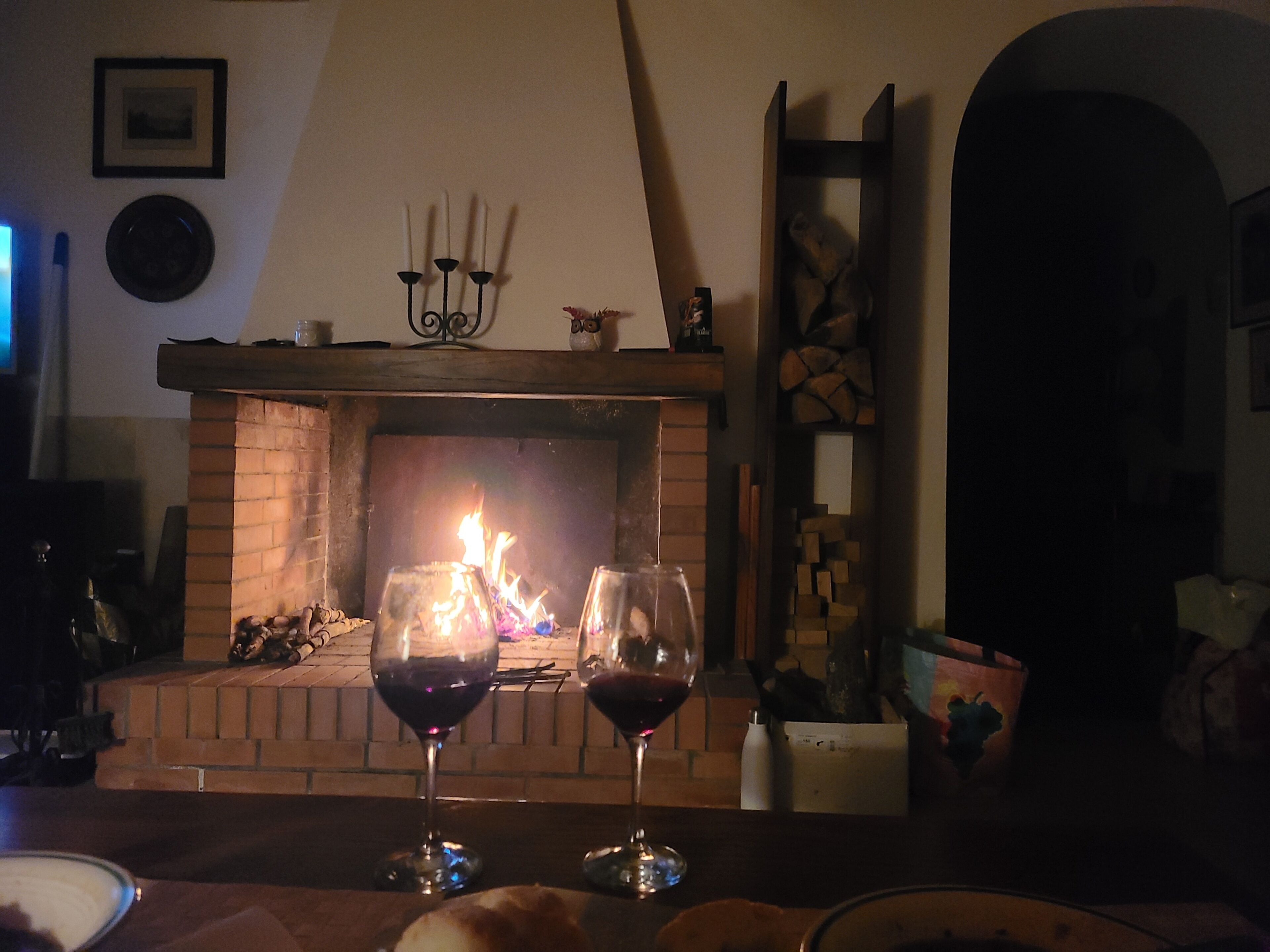 A fireplace with burning logs, two glasses of red wine, and a brick mantelpiece.