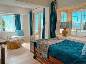 A bedroom with a bed, a hammock, a wooden chest of drawers, a potted plant, and a view of the outdoors.