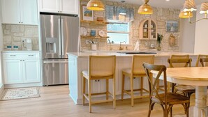 Une cuisine moderne avec des armoires blanches, un réfrigérateur en acier inoxydable, un mur décoratif en pierre et un coin repas avec des chaises en bois et une table ronde.