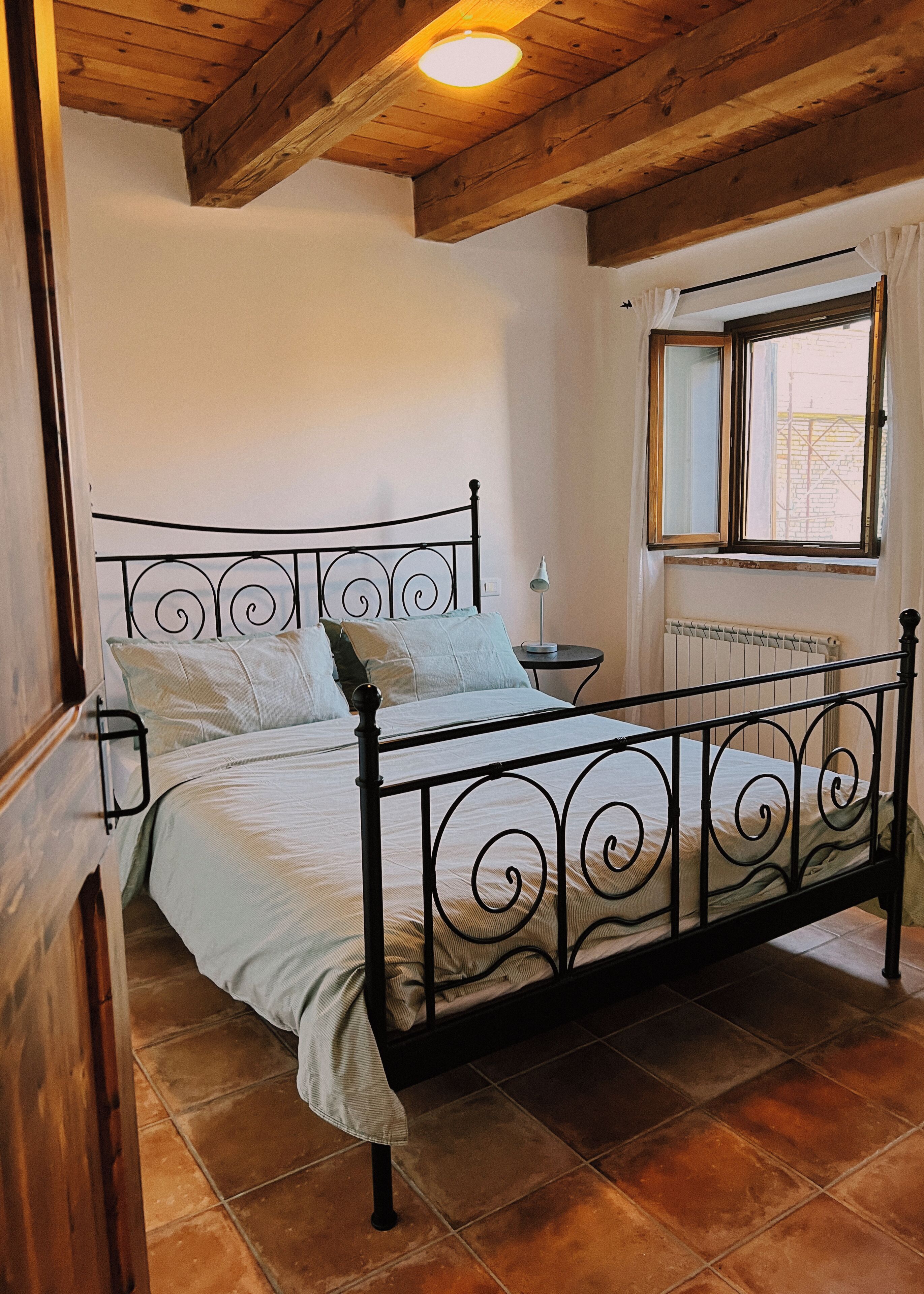 A bedroom with a metal bed frame, a bedside table, a lamp, and a window with a view.