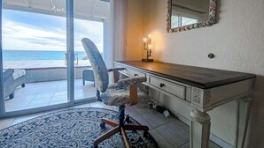 Quarto com uma grande porta de correr de vidro que dá para uma varanda com vista para o oceano. Há uma escrivaninha com gavetas e um espelho na parede.