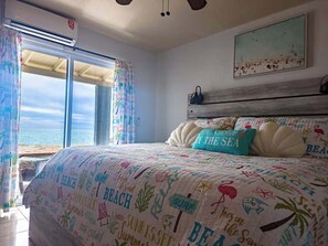 A bedroom with a beach-themed bedspread, a view of the sea through a large window, and a wooden headboard. - Once Upon a Tide | Oceanfront + Sleeps 8 (Playa Encanto)