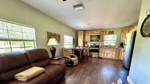 A living room with a brown leather sofa, a wooden coffee table, and a dining area with a table and chairs. The kitchen features wooden cabinets, a refrigerator, and an oven.