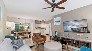 Ein modernes Wohnzimmer mit Deckenventilator, Essbereich, K üche und großem Fernseher an der Wand.