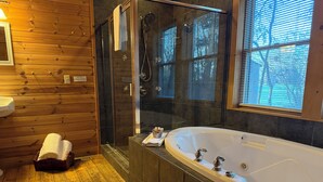 Une salle de bain avec un plancher en bois, une douche en verre, une grande baignoire et une fenêtre avec des stores.