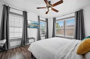 Ein Schlafzimmer mit einem Bett, einem Deckenventilator, einem Fenster mit Jalousien und einem an der Wand montierten Fernseher.