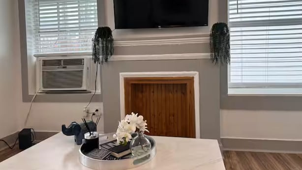 Un salon doté d’un téléviseur à écran plat fixé au-dessus d’une cheminée, d’une table basse en marbre agrémentée d’objets décoratifs et d’une grande fenêtre avec des stores.