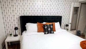 Un lit bien fait, avec une tête de lit noire, des oreillers blancs et orange, et une table de chevet blanche sur laquelle se trouve une lampe.