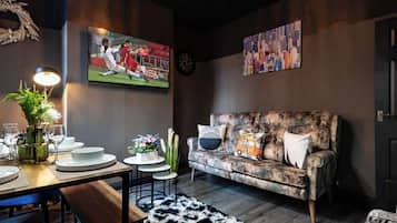 Ett modernt vardagsrum med en soffa, ett matbord dukat med tallrikar och glas, en tv och en tavla som föreställer en stadsbild.
