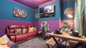Een woonkamer met een rode bank, een eettafel met borden en glazen, een aan de muur bevestigde televisie, en een decoratieve krans.