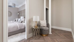 Ein Schlafzimmer mit einem Bett, einem Nachttisch mit Lampe, einem Sessel und einem Flur, der in einen anderen Raum führt.