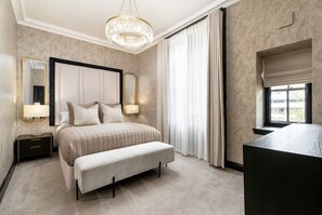 A bedroom with a large bed, bedside lamps, a bench, a chandelier, a window with curtains, and a mirror. - Luxury City Centre Apartments  (Edinburgh)