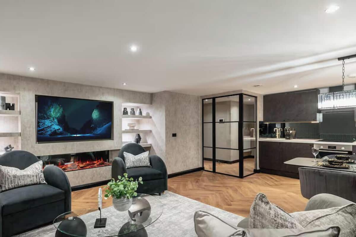 A modern living room with a flat-screen TV, two armchairs, a coffee table, and a fireplace.