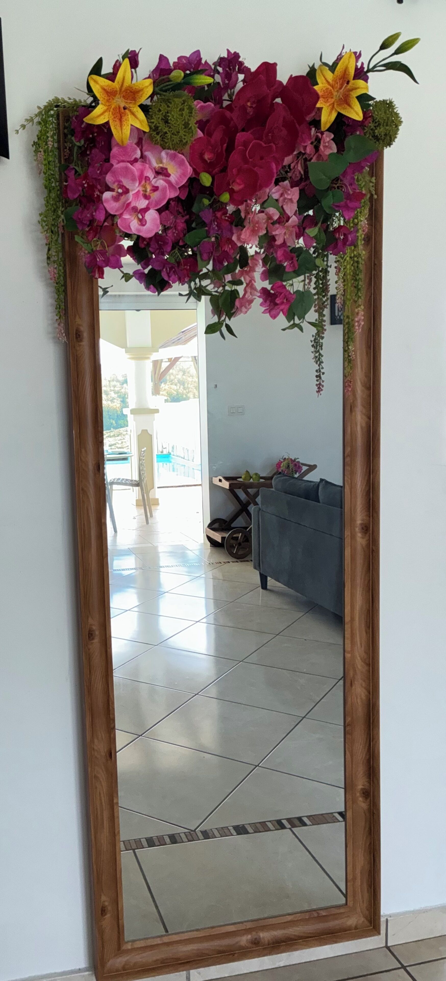 Un miroir surmonté d’un arrangement floral, qui reflète une pièce avec un canapé gris et un sol carrelé.