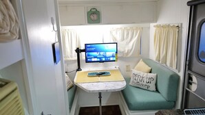 Interior compacto de una caravana con kitchenette, una pequeña mesa con una computadora, un sofá color turquesa y un cojín con texto.