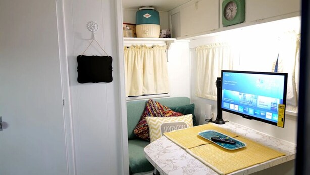 Un espacio habitable pequeño con una cama, una mesita con un monitor de computadora y una ventana con cortinas.