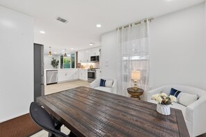 A modern kitchen with white cabinets, a dining area with a wooden table, and a white sofa with a vase of flowers. - Casa Esmeralda -Heated Pool, Luxury BBQ & Patio (Houston)