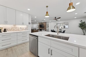 A modern kitchen with white cabinetry, a stainless steel dishwasher, a large island with a sink, pendant lighting, and a ceiling fan. - Casa Esmeralda -Heated Pool, Luxury BBQ & Patio (Houston)