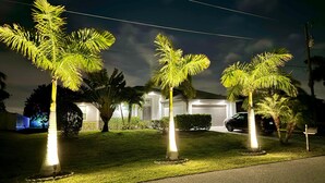 Un barrio residencial con un césped bien iluminado, palmeras, un garaje y un coche aparcado.