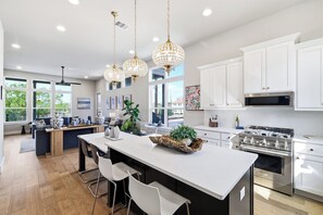 A modern kitchen with white cabinets, a large island with bar stools, pendant lighting, and a stainless steel stove. - The Carlisle - A Birdy Vacation Rental (San Antonio)