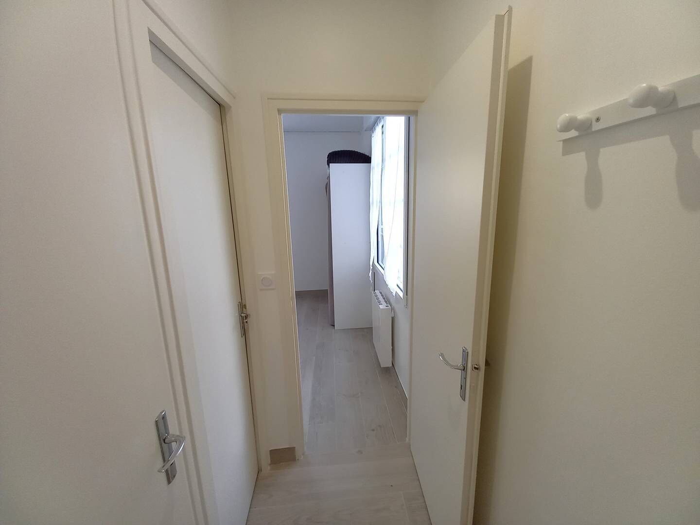 A narrow hallway with white doors, a radiator, and a window with curtains.