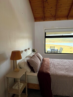 Een slaapkamer met een bed, een nachtkastje met een lamp, een raam met uitzicht op een landschap en een houten plafond.