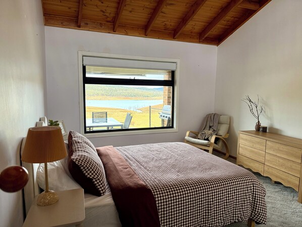 Une chambre avec une grande fenĂȘtre, un lit, une chaise, une commode et une lampe.