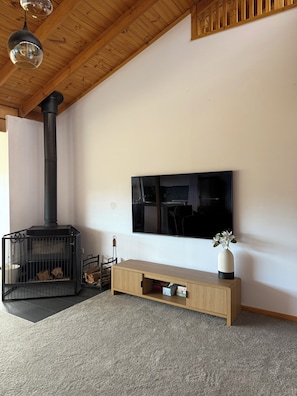 Een woonkamer met een zwart houtkacheltje, een flatscreen-televisie, een houten bankje en een vaas met bloemen.