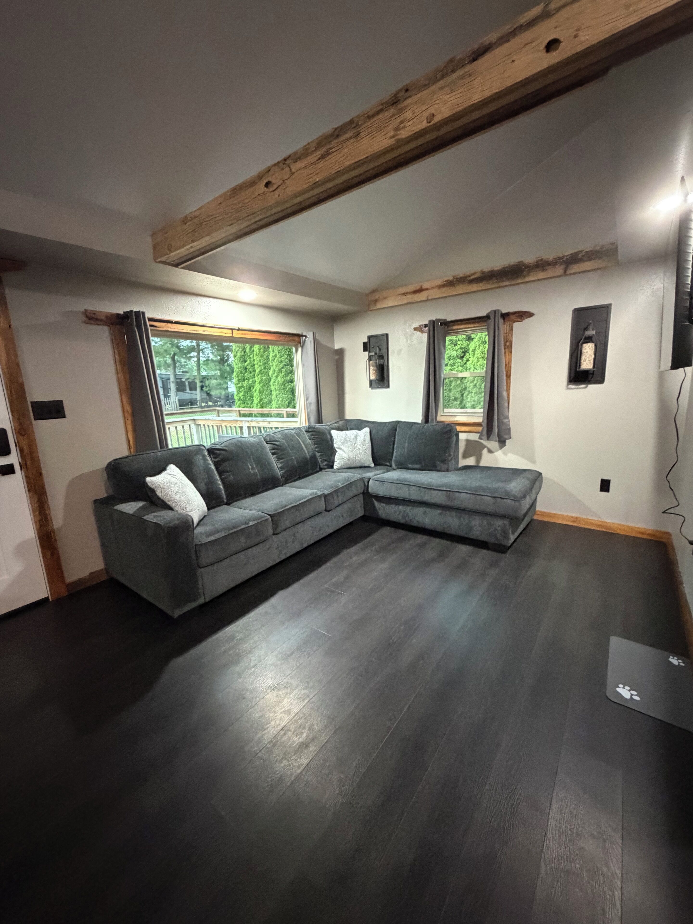 Una habitación con un sofá seccional gris, vigas de madera en el techo y una ventana con vista a follaje.