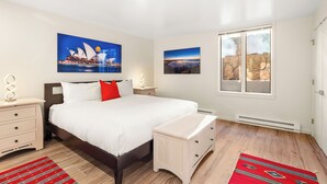 Un dormitorio con cama, mesitas de noche, cómoda, ventana con vistas y dos cuadros decorativos.