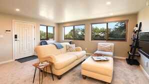 A living room with a beige sofa, ottoman, and a small wooden side table. There are two windows providing natural light and a door with a doormat. - SF Bay Area: Private Luxury “Treehouse” Retreat (Pleasant Hill)
