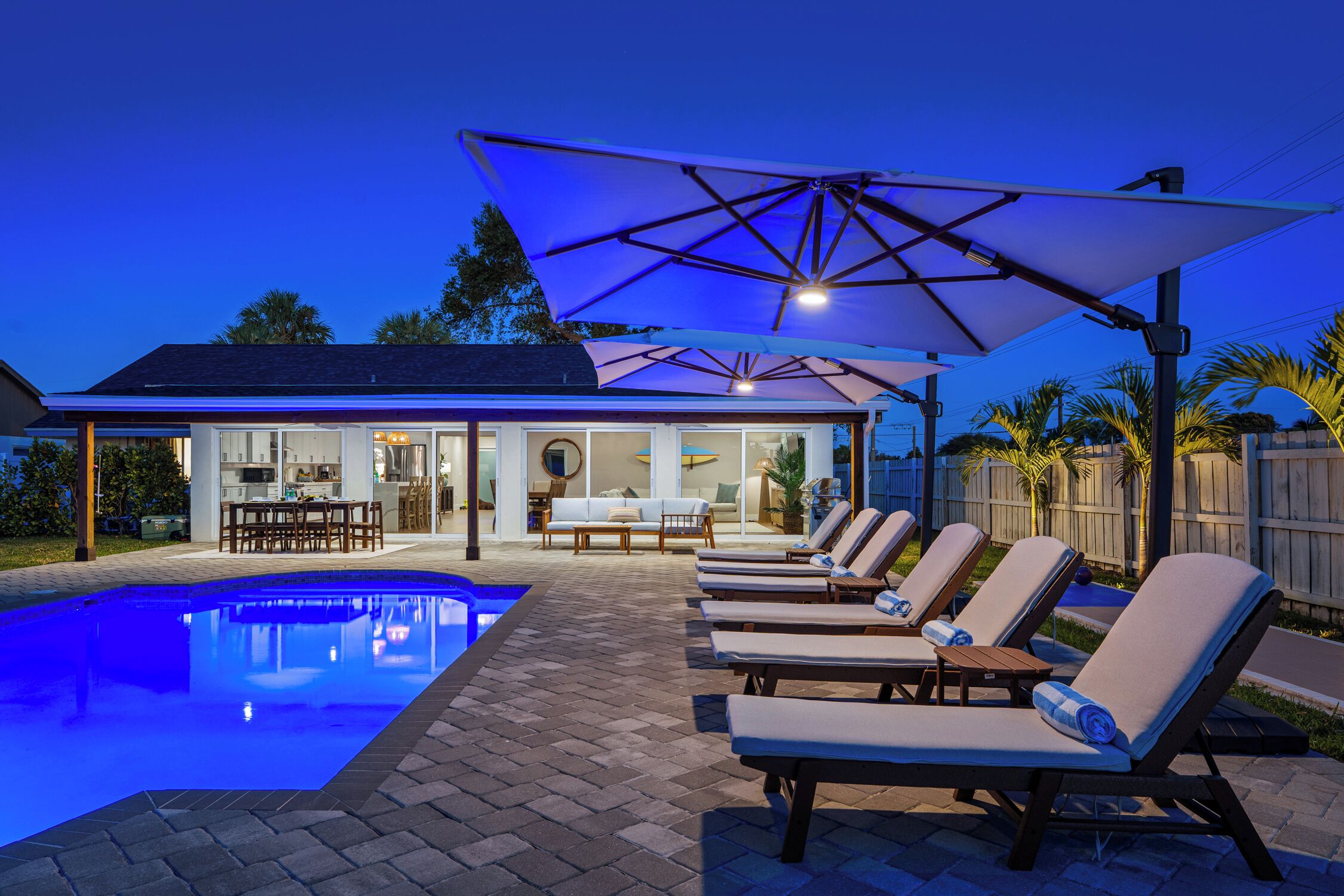 Una piscina con sillas de descanso y sombrillas, un patio cubierto y vista a una casa con comedor.