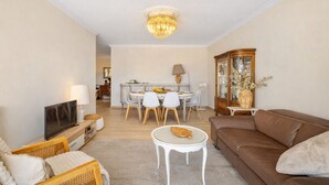 Un salon avec un canapé marron, un téléviseur, une table à manger avec des chaises, un lustre et un meuble avec des portes en verre.