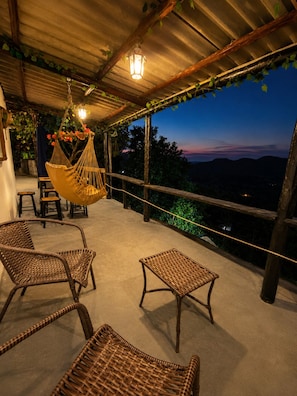 A covered outdoor seating area with wicker furniture and a hanging chair, overlooking a mountainous landscape.
