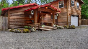 En stuga av trä med en veranda, ett garage och en grusuppfart.