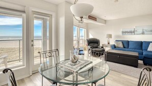A living room with a glass table, a blue sofa, a leather chair, and a view of the beach through large windows.