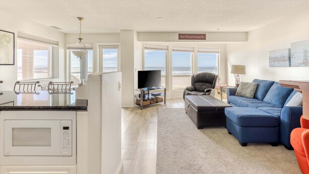 A modern living room with a sectional sofa, a flat-screen TV, a dining area with a microwave, and large windows offering a view.