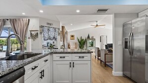 A modern kitchen with granite countertops, white cabinetry, a stainless steel refrigerator, and a ceiling fan. - The Salty Peacock near Englewood Beach (Rotonda West)