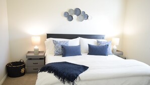 Ein ordentlich gemachtes Bett mit weißer und blauer Bettwäsche, zwei Nachttische mit Lampen, ein Wandbild und ein Korb aus Weidengeflecht.