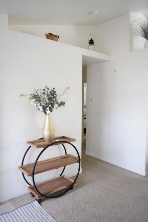Un pasillo con pared blanca, una estantería de madera con un jarrón de flores y un farol negro en la pared.
