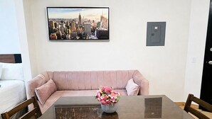 A dining area with a glass-top table, wooden chairs, a pink sofa, and a cityscape painting on the wall. - CABO DOWNTOWN STUDIOS: 1BR Studio 2 doubles perfect location close to marina #3 (Cabo San Lucas)