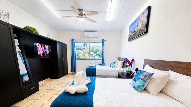 A hotel room with a bed, a TV, a ceiling fan, a window with curtains, and a painting on the wall. - CABO DOWNTOWN STUDIOS: 1BR Studio 2 doubles perfect location close to marina #3 (Cabo San Lucas)