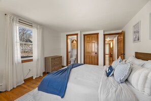 Een slaapkamer met een groot bed, een dressoir, een raam met gordijnen en een deur naar een andere kamer.