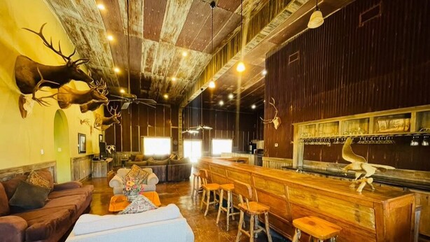 Un intérieur rustique avec un coin bar, des tabourets en bois, un canapé et des têtes d’animaux fixées au mur.