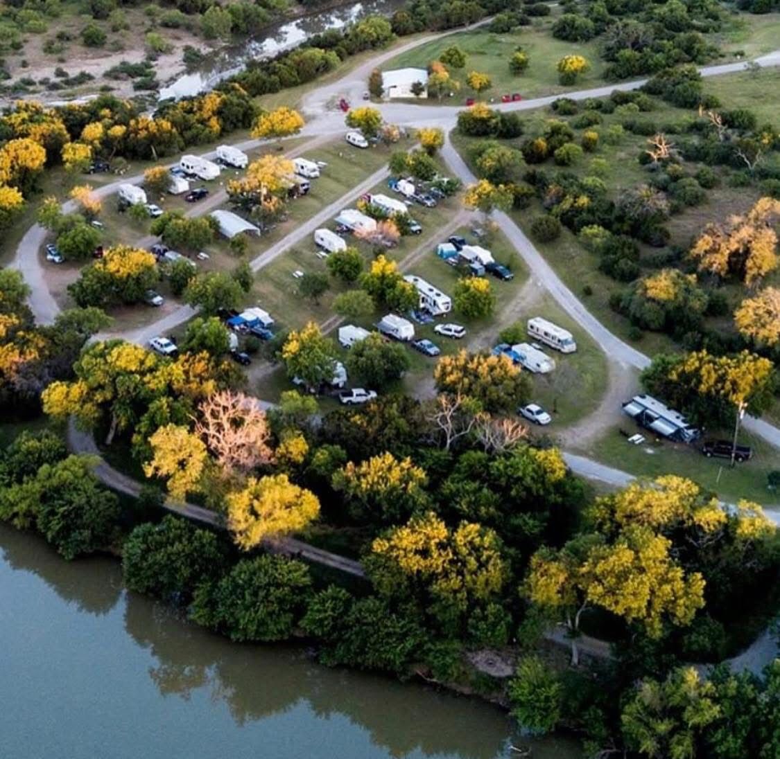 Vista aérea de un camping con vehículos recreativos y tiendas de campaña, rodeado de árboles y una masa de agua.