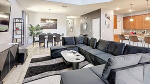 Ein modernes Wohnzimmer mit einer Eckcouch, einem Essbereich mit Tisch und Stühlen sowie einer Küche mit Holzausstattung und Pendelleuchten.