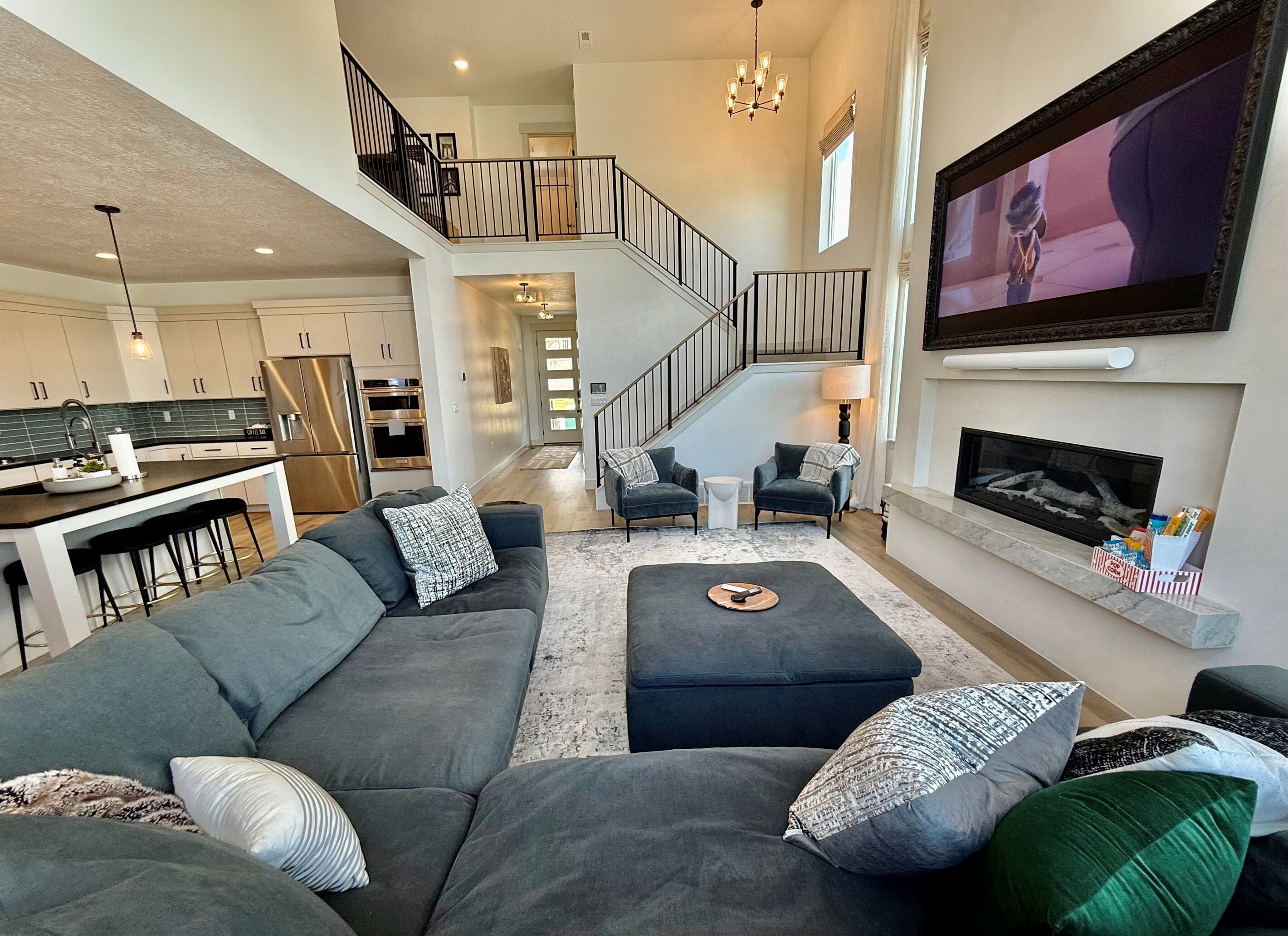 A spacious living area with a large gray sofa, a fireplace, a TV mounted on the wall, and a kitchen in the background.